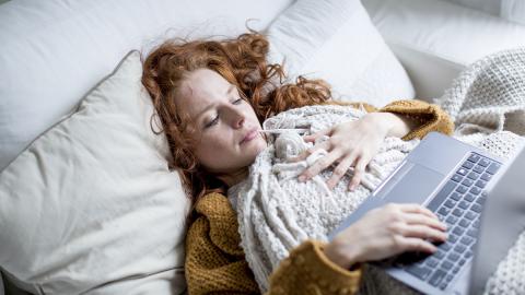 Frau liegt im Bett mit Laptop auf der Decke.